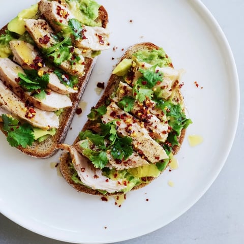 Golden grilled chicken slices layered over creamy smashed avocado on crispy sourdough toast, topped with chili flakes and honey.  